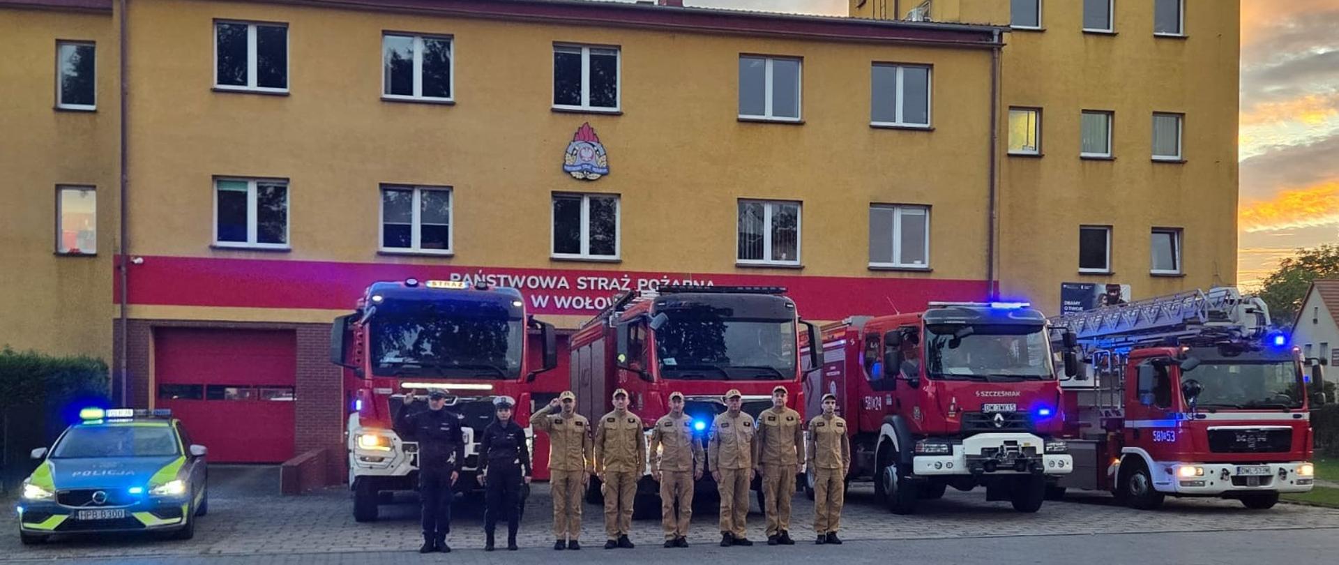 Strażacy oraz policjanci pozują do zdjęcia na tle komendy Państwowej Straży Pożarnej w Wołowie wraz ze swoimi wozami bojowymi i radiowozem z włączonymi sygnałami świetlnymi.