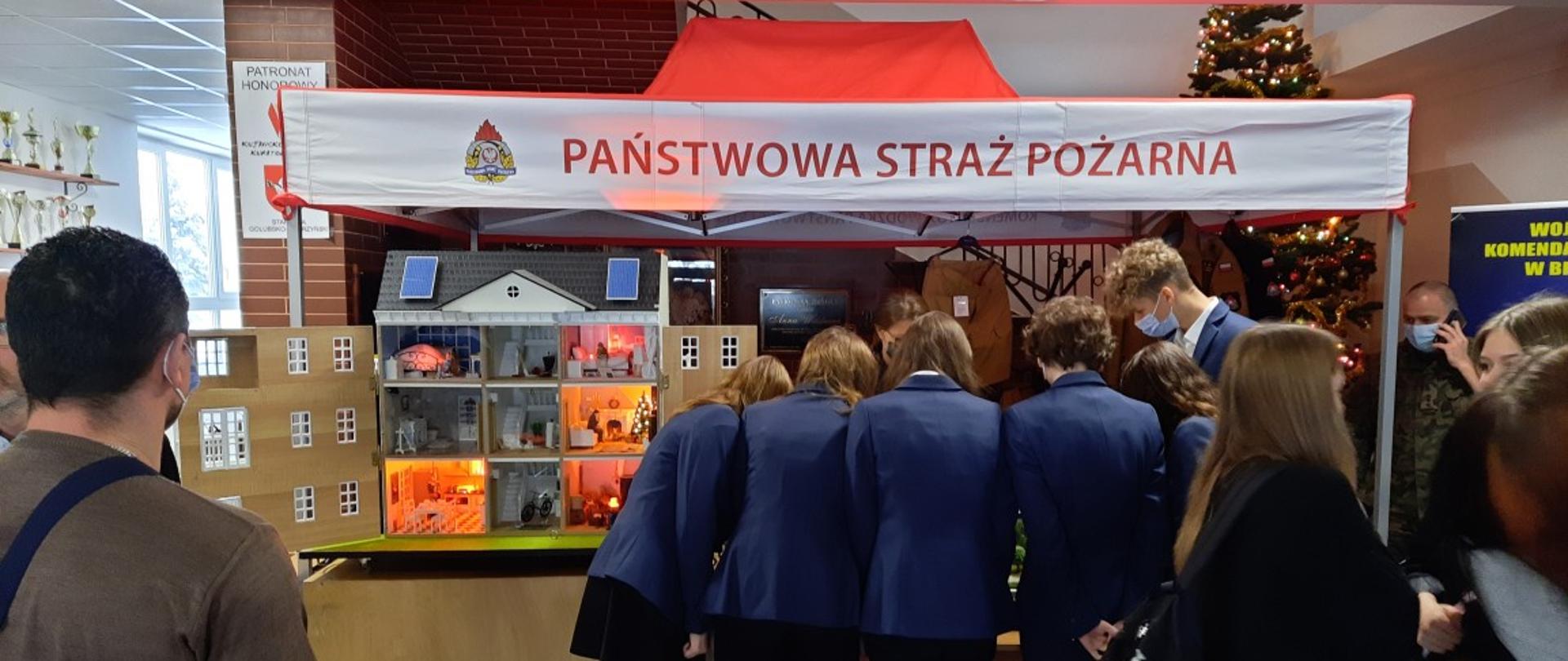 Widok na uczestników Olimpiady, którzy odwiedzają stoisko PSP. Stoją tyłem, zainteresowani są makietą. Ubrani są w szkolne mundurki, reprezentujące szkołę. W ramach stoiska widoczne symulatory zagrożeń.