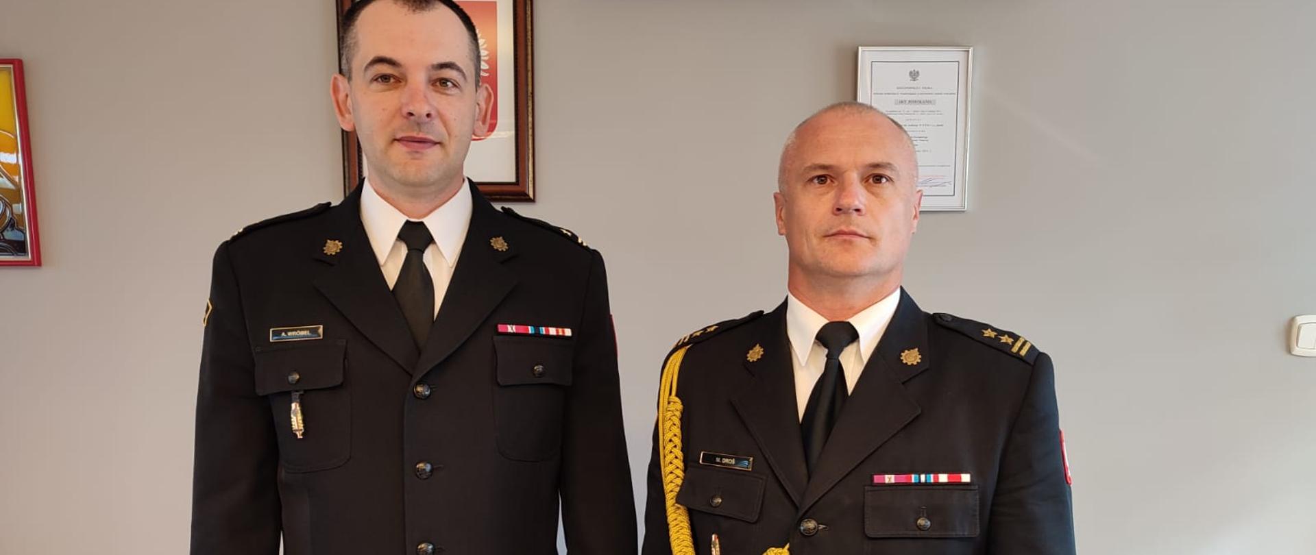 Dwóch strażaków stoi w mundurach wyjściowych w tle ściana pomieszczenia.