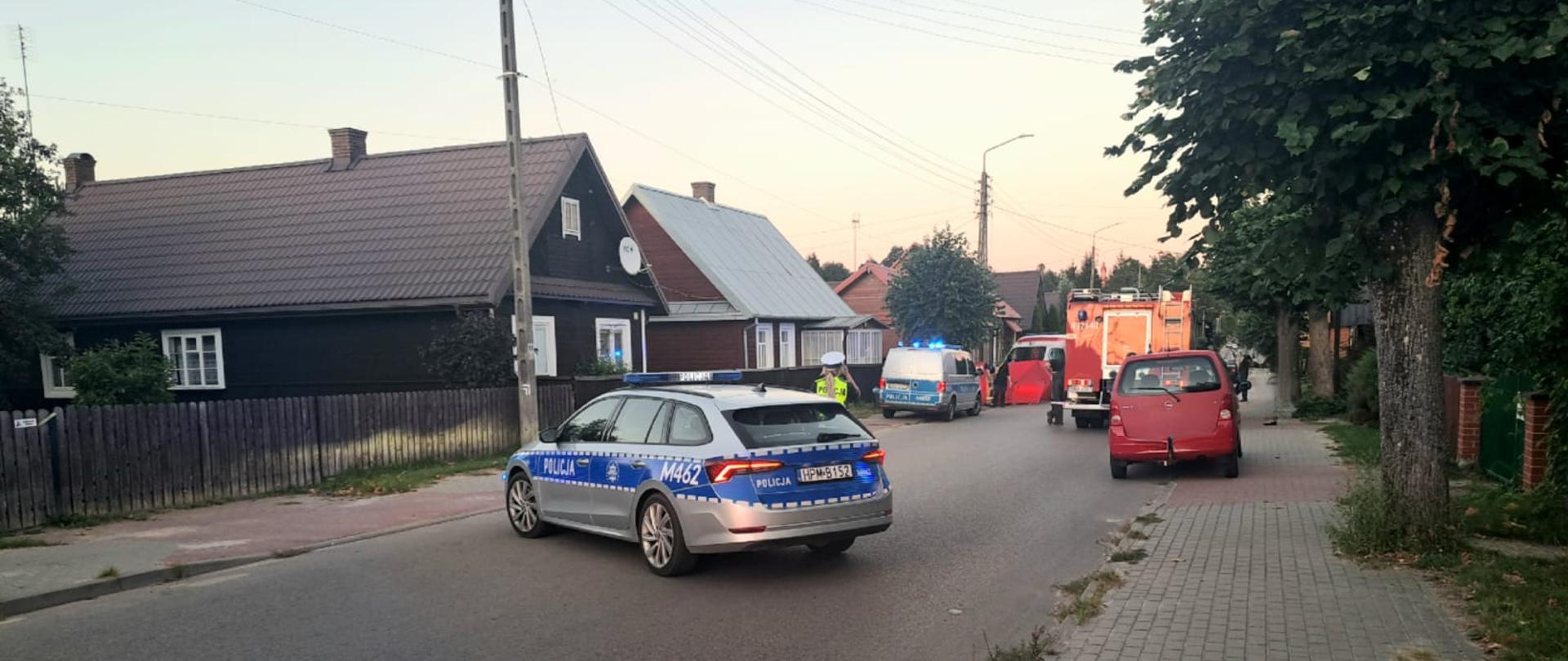 Na środku drogi w zabudowie wiejskiej stoją dwa samochody Policji i jeden czerwony strażacki wóz bojowy.