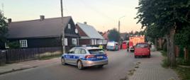 Na środku drogi w zabudowie wiejskiej stoją dwa samochody Policji i jeden czerwony strażacki wóz bojowy.