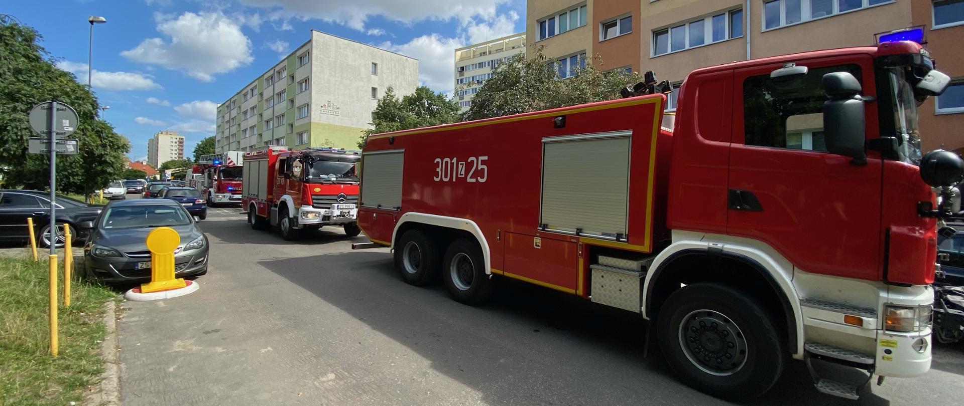 Na pierwszym panie dwa specjalistyczne samochody ratowniczo gaśnicze. Na drugim planie budynek mieszkalny wielorodzinny oraz samochód straży pożarnej z drabina mechaniczną.