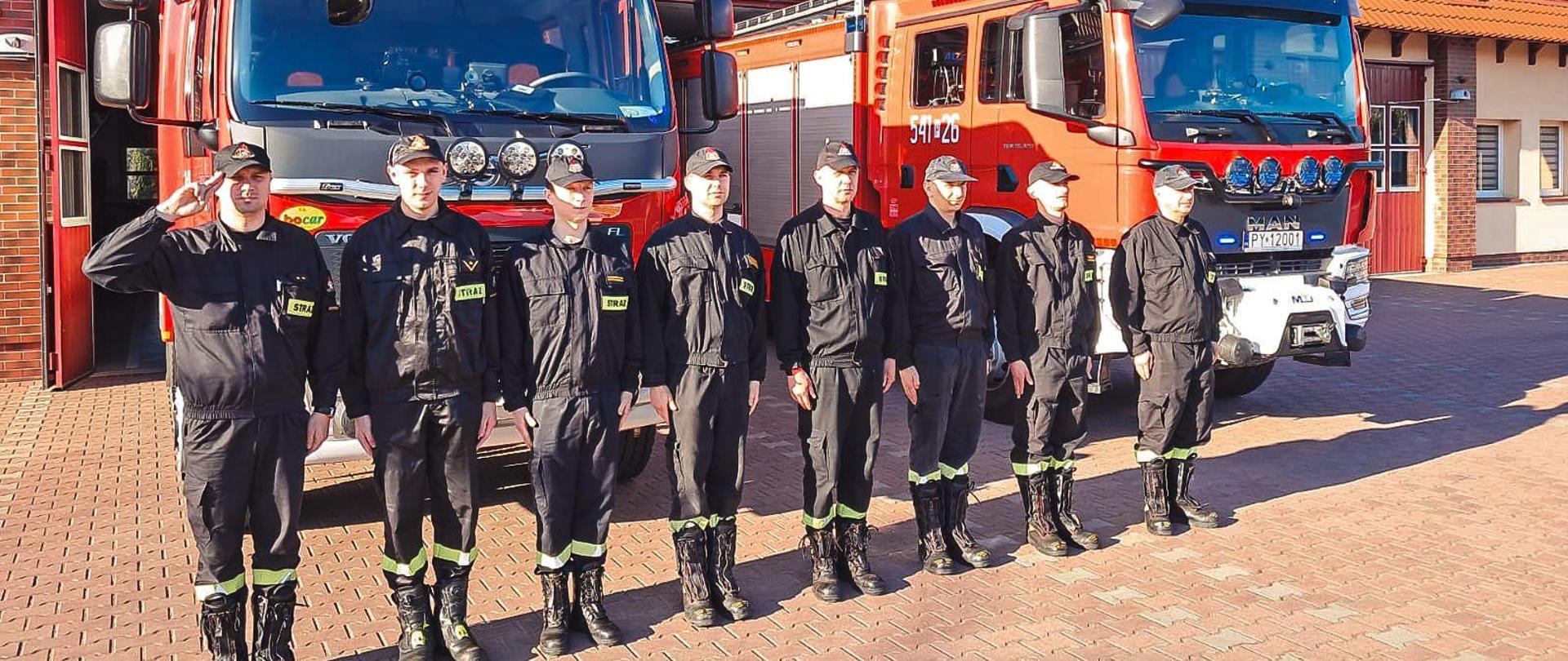 Strażacy w ubraniu koszarowym stoją na placu manewrowym przed budynkiem strażnicy oraz wozami strażackimi.