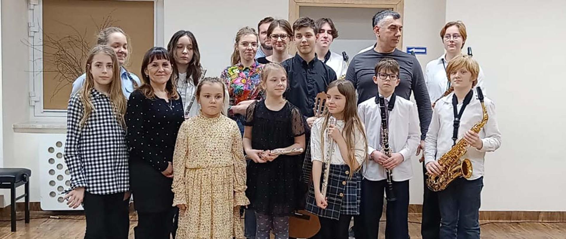 Na zdjęciu grupa kilkunastu osób, dorośli i młodzież, stoją w pomieszczeniu twarzami zwróconymi do robiącego zdjęcie, kilka osób trzyma w rękach instrumenty muzyczne - saksofon, klarnet i flety.