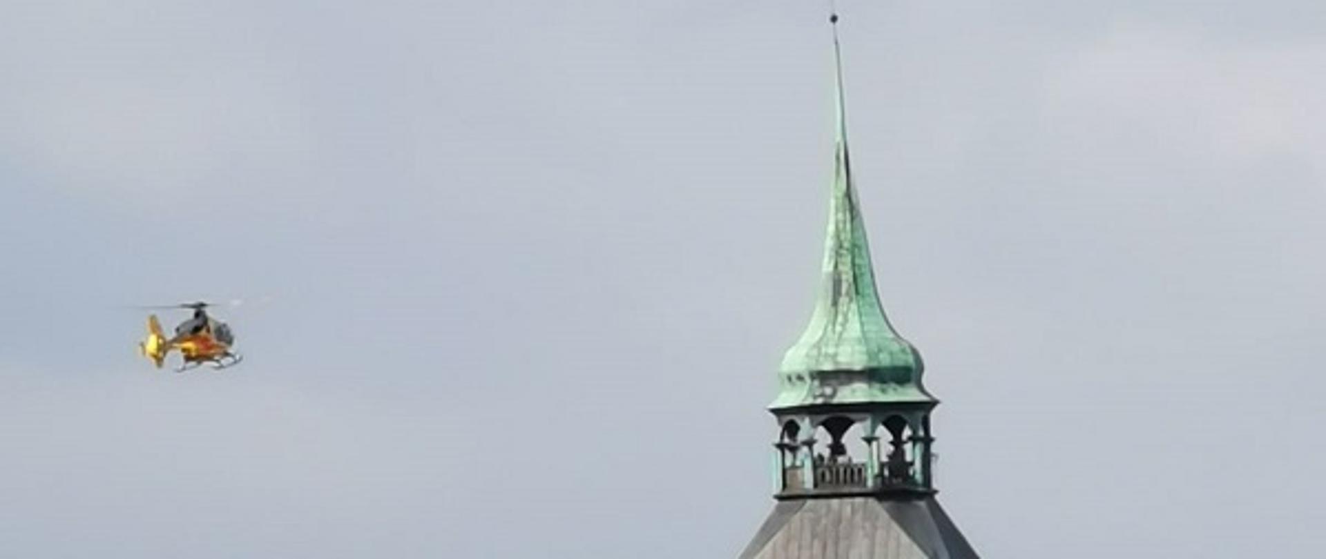 Zdjęcie przedstawia lądujący przy kościele mariackim w Białogardzie śmigłowiec Lotniczego Pogotowia Ratowniczego. Żółty śmigłowiec unosi się nad dachami budynków mieszkalnych