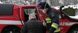 Na zdjęciu strażak pomaga starszej osobie przejść z ośrodka szczepień do samochodu oznakowanego. W tle widzimy samochód oznakowany oraz drzewa pokryte śniegiem.
