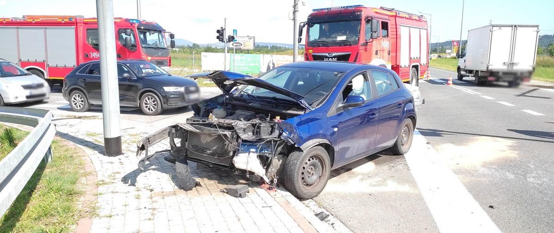 Samochód z rozbitym przodem częściowo na chodniku a częściowo na jezdni. W tle samochody pożarnicze.