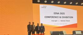 Czterech mężczyzn pozuje do wspólnego zdjęcia na tle dużego ekranu z logiem konferencji EENA 2025 i napisem „Conference & Exhibition – 9-11 April, Helsinki, Finland”; na pierwszym planie duży napis Eena2025.