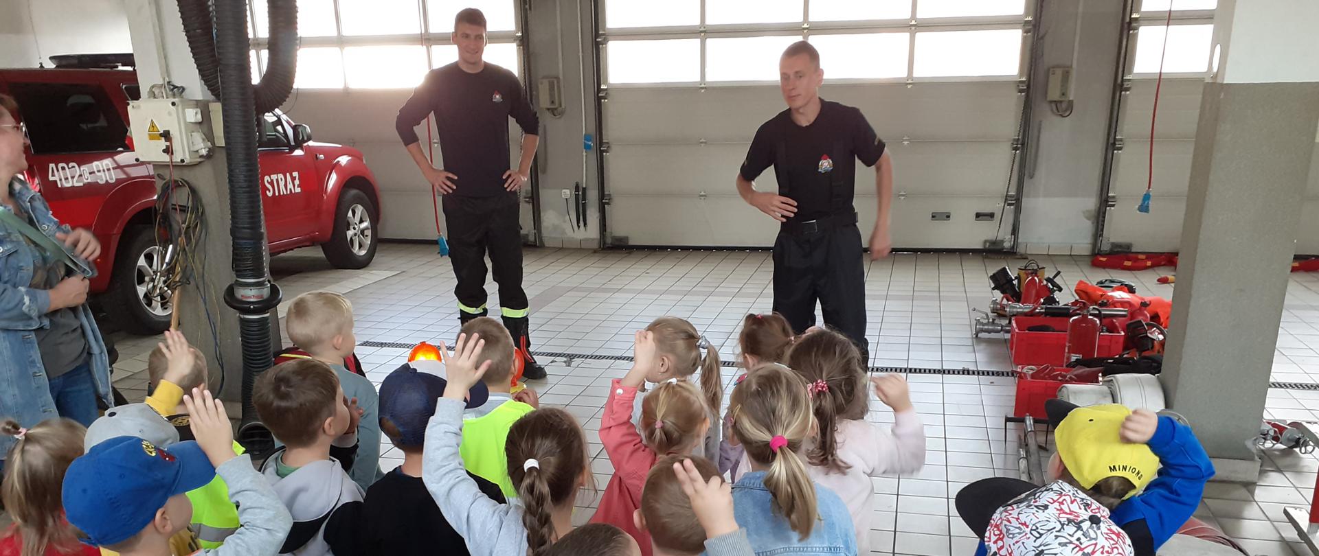 Na zdjęciu widać grupę przedszkolaków biorących udział w pogadance z dwoma strażakami. Dzieci unoszą ręce w odpowiedzi na zadane przez strażaka pytanie. Pogadanka odbywa się wewnątrz garażu Jednostki Ratowniczo-Gaśniczej. W tle widać bramy garażowe oraz pożarniczy samochód terenowy i rozłożony sprzęt ratowniczo-gaśniczy.