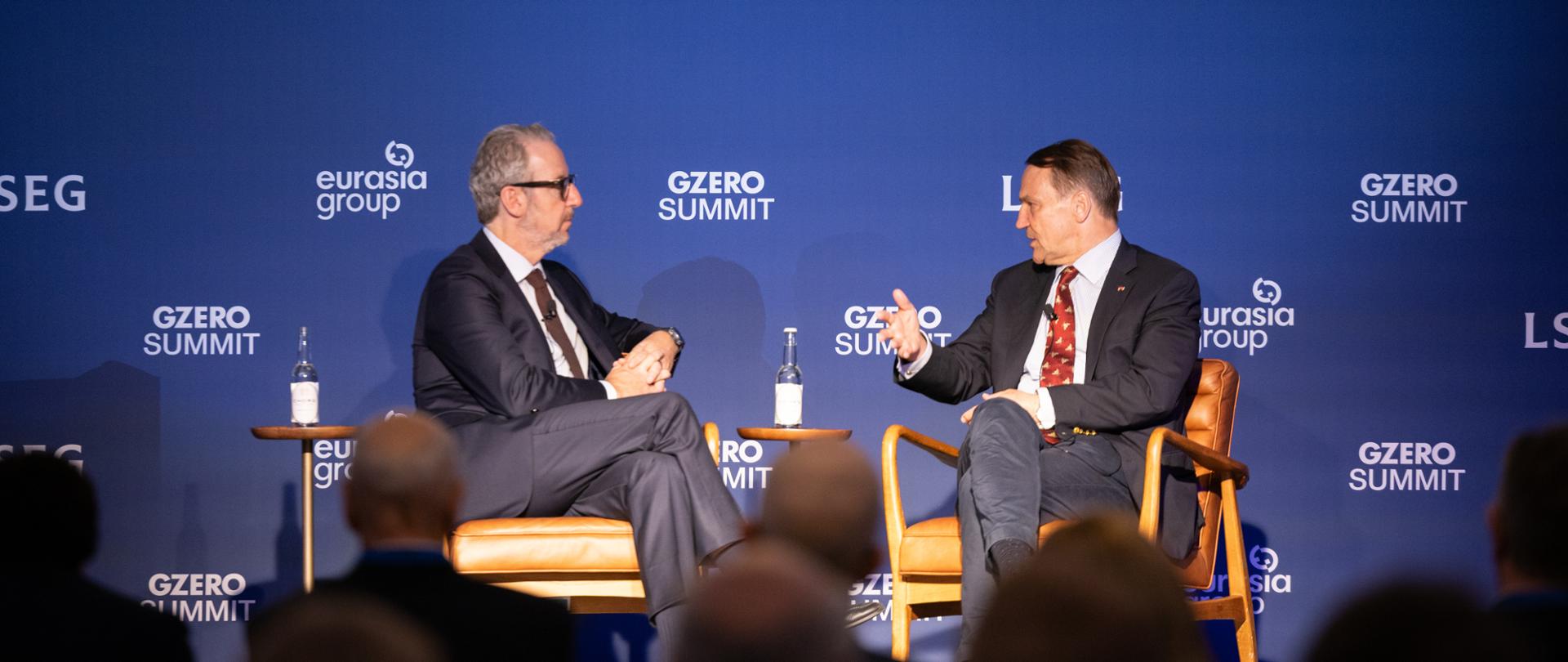 Moderator and Deputy Prime Minister Sikorski are seated on stage. Behind them is a blue backdrop displaying “GZERO SUMMIT London 2026” and the logos of the event’s organisers and partners.
