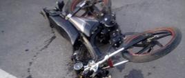 Zderzenie auta osobowego z motorowerem. Droga powiatowa. Na jezdni leży motorower uczestniczący w wypadku. Wokół niego plamy płynów eksploatacyjnych. 