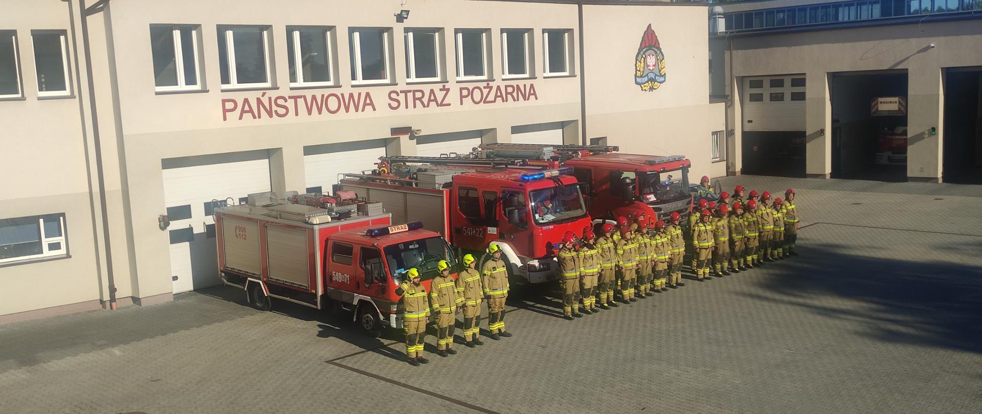 Przed garażami JRG Tczew stoją samochody ratowniczo gaśnicze z włączonymi światłami alarmowymi, przed pojazdami 27 strażaków w pełnym umundurowaniu specjalnym oddaje honor.