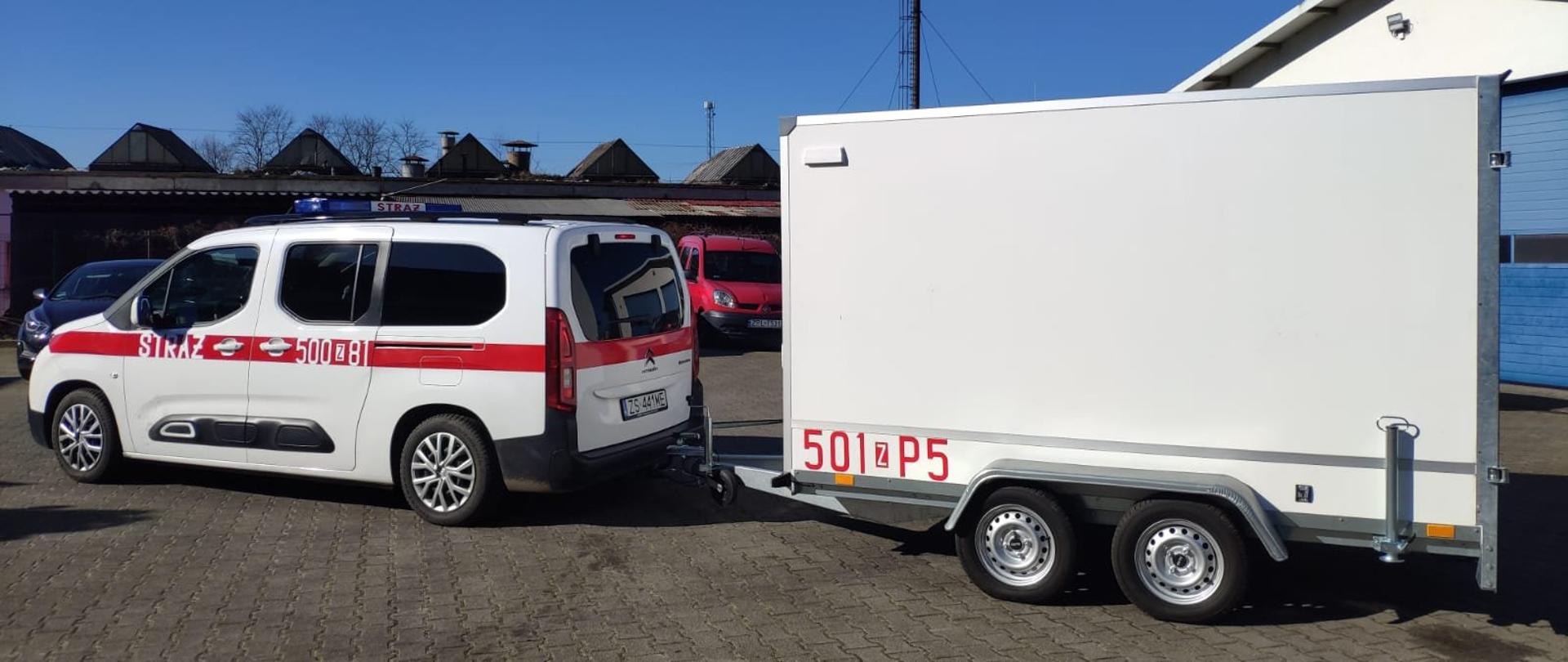 Na zdjęciu widoczny plac z nawierzchnia z polbruku. Widoczny lewy bok stojącego białego mikrobusu z czerwonym pasem przebiegającym wzdłuż pojazdu z białym napisem straż i numerem 500 z 81, do którego przyłączono białą dwuosiową przyczepę z czerwonym napisem na dole skrzyni 501 z P5. W tle widoczne fragmenty zabudowań warsztatowych. Niebo pogodne, błękitne.