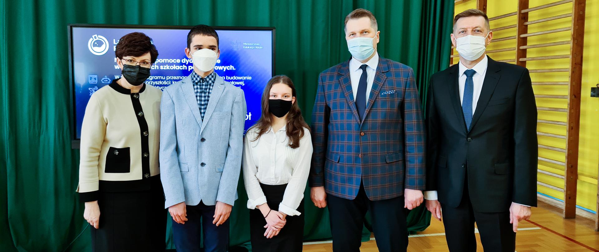 Minister Przemysław Czarnek, Lubelska Kurator Oświaty Teresa Misiuk, dyrektor szkoły Mirosław Wójcik oraz dwoje uczniów szkoły pozuje do zdjęcia na sali gimnastycznej.