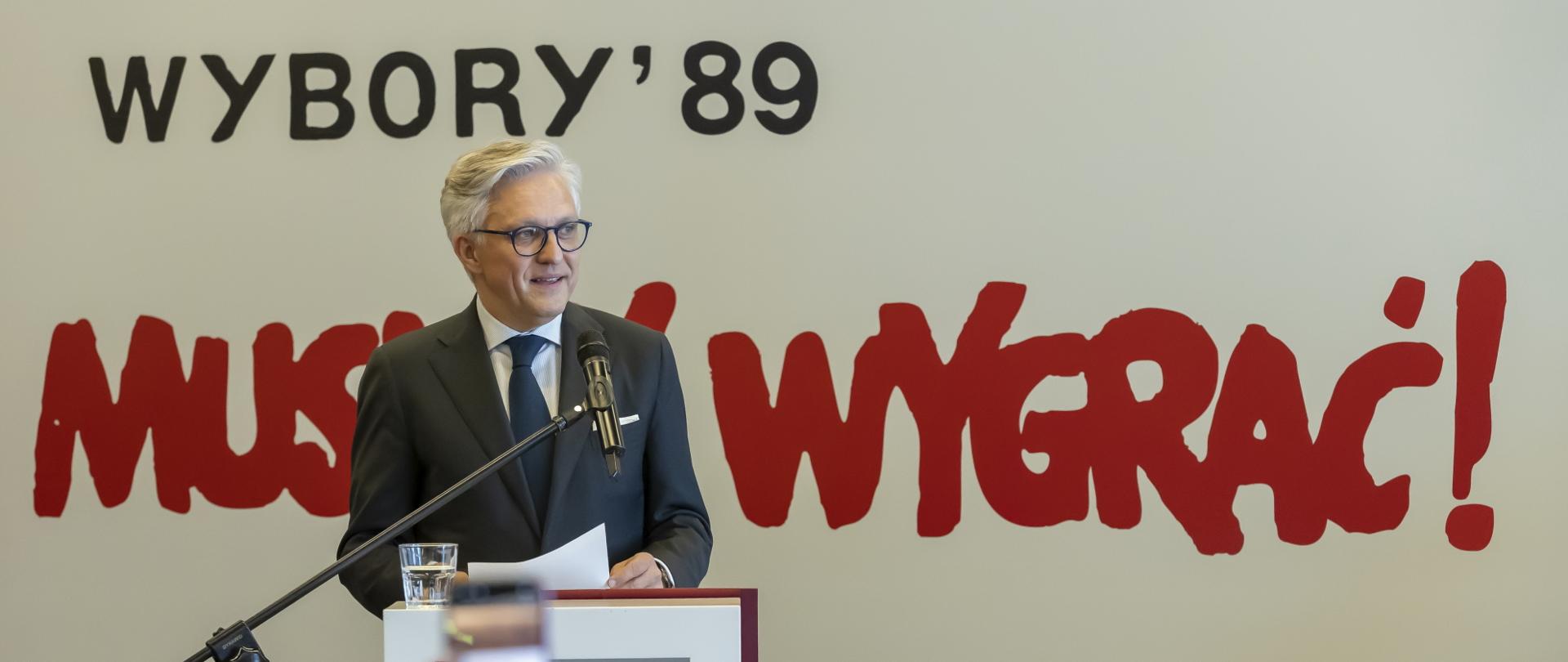 Podsekretarz Stanu Artur Harazim w ciemnym garniturze, białej koszuli i okularach stoi przed publicznością na scenie. Za nim napis 'Wybory '89. Musimy wygrać!"
