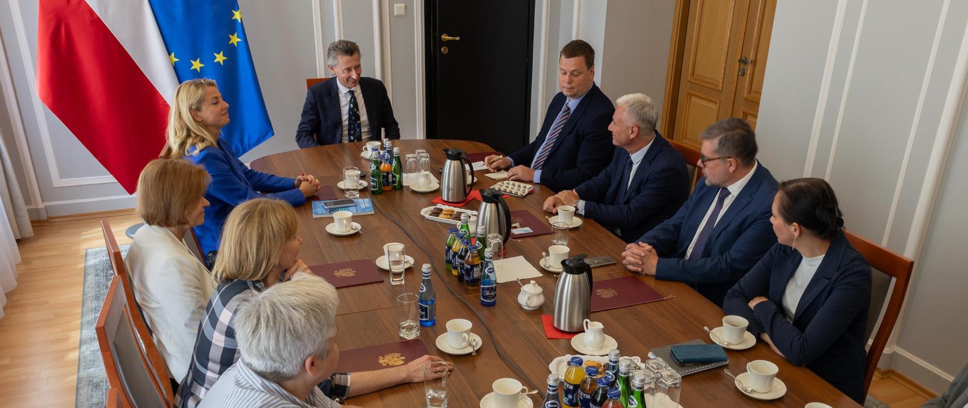 Widok na małą salę, wokół owalnego stołu siedzą ludzie, za nimi pod ścianą flagi Polski i UE.