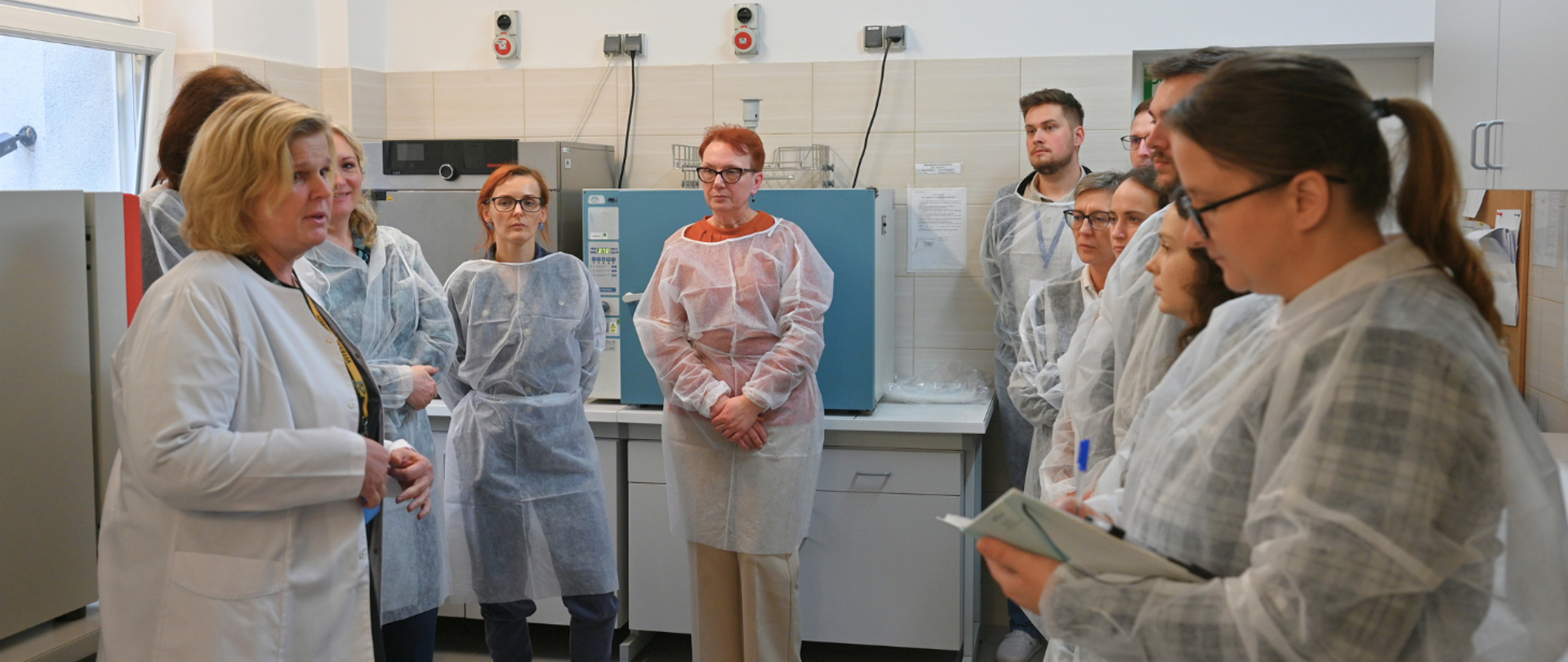 Grupa osób w jednorazowych fartuchach ochronnych stoi w laboratorium, słuchając kobiety w białym kitlu, która prowadzi omówienie lub szkolenie. W tle widać sprzęt laboratoryjny oraz szafki.