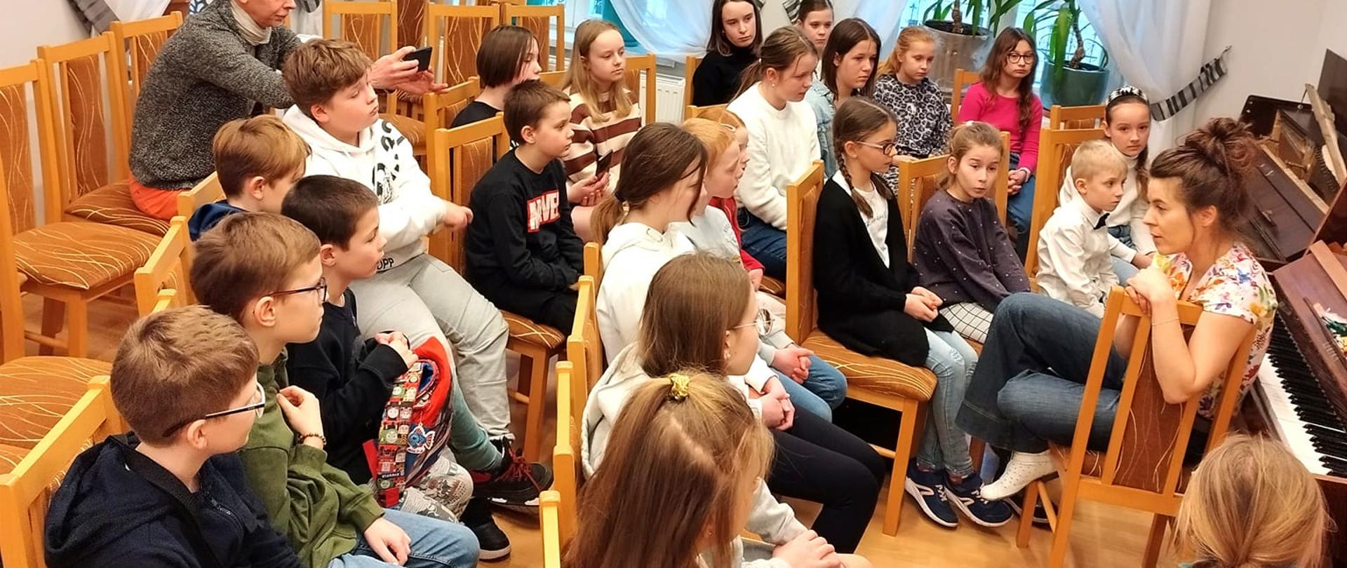 Zdjęcie zrobione w auli szkoły muzycznej przedstawia pania siedzącą na krześle prowadząca warsztaty z tyłu stoi pianino . Na krzesłach siedzą uczniowie oraz nauczyciel bioracy udział w warsztatach 