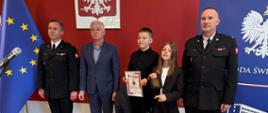 Na zdjęciu widoczny jest Świętokrzyski Komendant Wojewódzki Państowej straży pożarnej starszy brygadier Marcin Charuba oraz Komendant Powiatowy Państowej straży pożarnej w ostrowcu Świętokrzyski młodszy brygadier Marcin Jasiński wraz z młodymi bohaterami podczas odbioru podziekowań za ich bohaterski czyn.