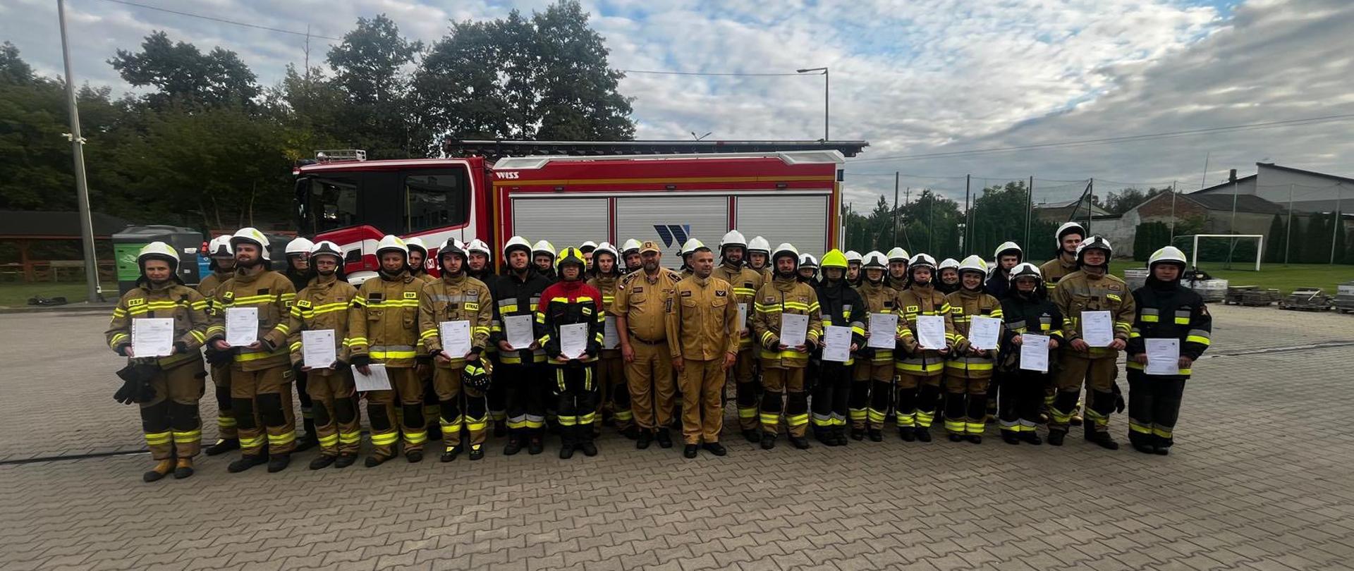 Duża grupa strażaków stoi razem przed czerwonym wozem strażackim. Wszyscy mają na sobie mundury bojowe i kaski, a część z nich trzyma w rękach certyfikaty. W środku grupy stoją także dwie osoby w jasnożółtych mundurach. Zdjęcie wygląda jak pamiątkowa fotografia po zakończonym szkoleniu lub ćwiczeniach.