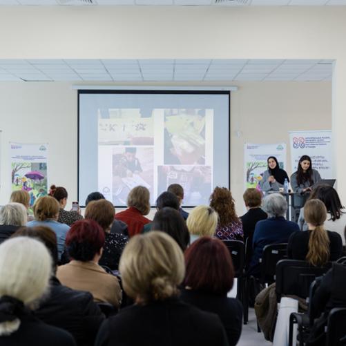 Sala konferencyjna z grupą osób, prezentują dwie kobiety, na prezentacji widać zdjęcia z warsztatów dla dzieci, w rogu flaga Gruzji