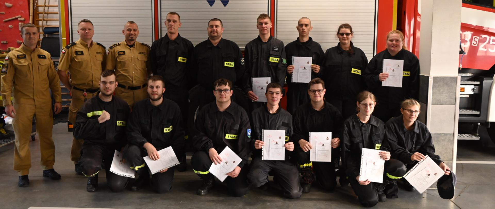 Grupa strażaków w ciemnych mundurach oraz 3 członków komisji Państwowej Straży Pożarnej pozuje do zdjęcia grupowego. W tle bok samochodu pożarniczego.