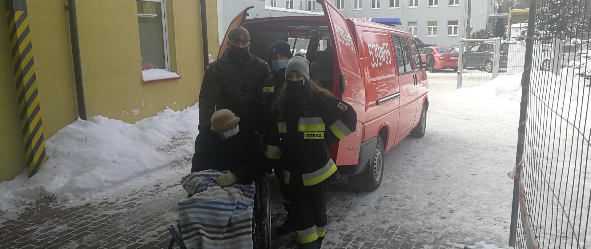 Starsza pani na wózku inwalidzkim, za nią stoją strażacy i żołnierz. W tle widać lekki samochód kwatermistrzowski z Ochotniczej Straży Pożarnej w Starych Jabłonkach
