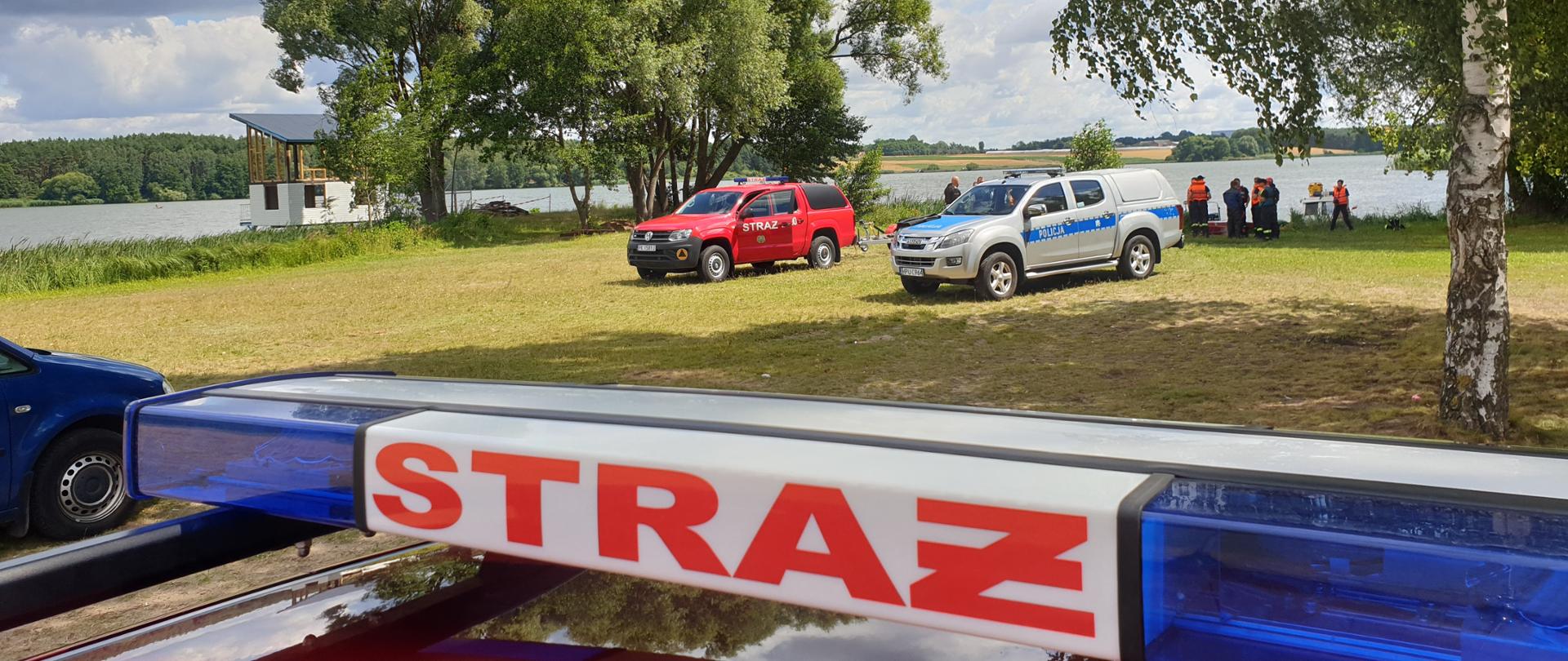 Zdjęcie przy zbiorniku. Widać pojazdy Policji i Jednostek Ochrony Przeciwpożarowej. Zdjęcie w ciągu dnia. Auta stoją na trawie. Występuje zachmurzenie. Widać ratowników, biorą udział w ćwiczeniach. W dalszej perspektywie zalew.
