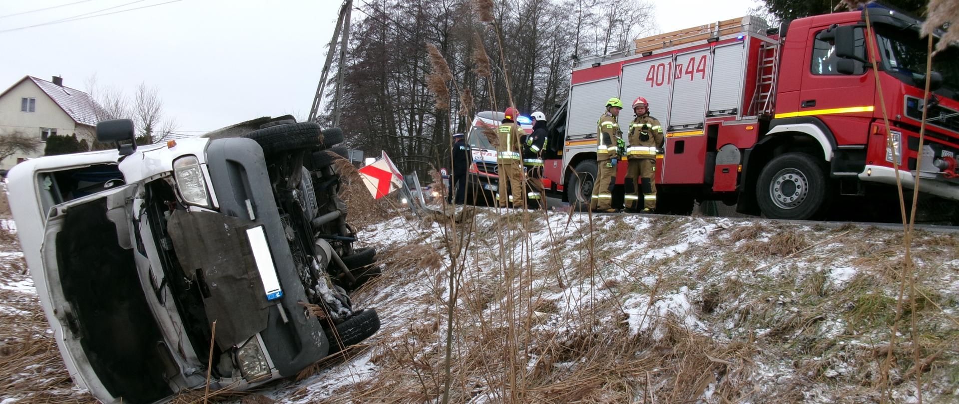 Zdjęcie przedstawia wypadek busa przewożącego pasażerów. Jest pora dzienna. Na zdjęciu widać wywrócony bus pasażerski na prawy bok i leży w rowie. Bus ma otwartą maskę przednią. Obok na jezdni stoją czerwony pojazdy pożarnicze a za nimi karetka Zespołu Ratownictwa Medycznego. Obok pojazdów stoją strażacy ubrani w ubrania specjalne w kolorze piaskowym na głowach maja hełmy. Przed pojazdem na ziemi leży akumulator