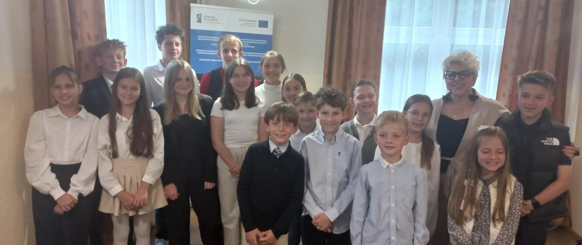 Sala kameralna siedemnastu uczniów klasy fortepianów stoją do wspólnego zdjęcia z swoją nauczycielką. 