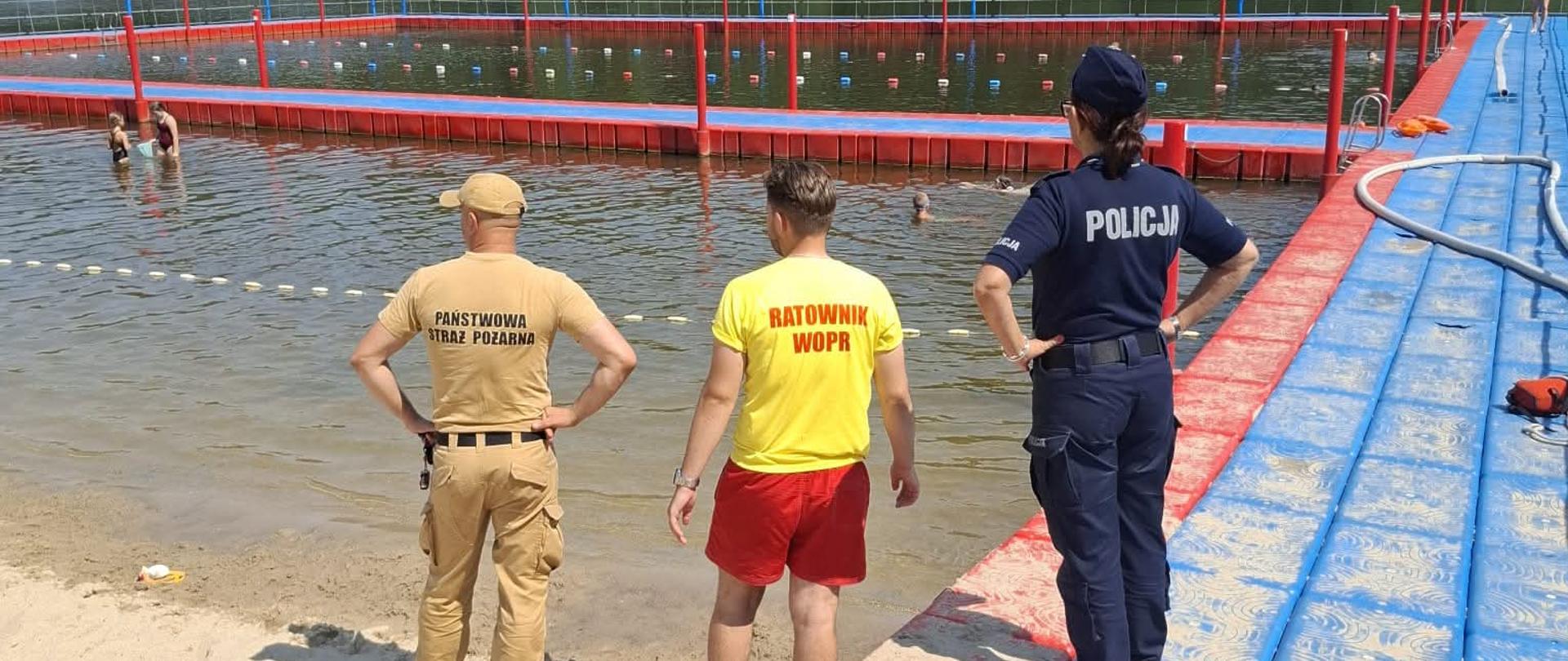 Widok z góry na strzeżone kąpielisko nad jeziorem lub zalewem w słoneczny dzień. Na pierwszym planie, na piaszczystej plaży, stoją trzy osoby odwrócone tyłem do obiektywu: funkcjonariusz Państwowej Straży Pożarnej w beżowym mundurze, ratownik WOPR w żółtej koszulce z napisem "RATOWNIK WOPR" i czerwonych spodenkach oraz policjantka w granatowym mundurze z napisem "POLICJA". Wszyscy obserwują wyznaczony bojami obszar kąpieliska, w którym widać kąpiących się ludzi. Kąpielisko otoczone jest pomostem z czerwonych i niebieskich elementów. W tle gęsty, zielony las.