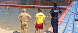Widok z góry na strzeżone kąpielisko nad jeziorem lub zalewem w słoneczny dzień. Na pierwszym planie, na piaszczystej plaży, stoją trzy osoby odwrócone tyłem do obiektywu: funkcjonariusz Państwowej Straży Pożarnej w beżowym mundurze, ratownik WOPR w żółtej koszulce z napisem "RATOWNIK WOPR" i czerwonych spodenkach oraz policjantka w granatowym mundurze z napisem "POLICJA". Wszyscy obserwują wyznaczony bojami obszar kąpieliska, w którym widać kąpiących się ludzi. Kąpielisko otoczone jest pomostem z czerwonych i niebieskich elementów. W tle gęsty, zielony las.