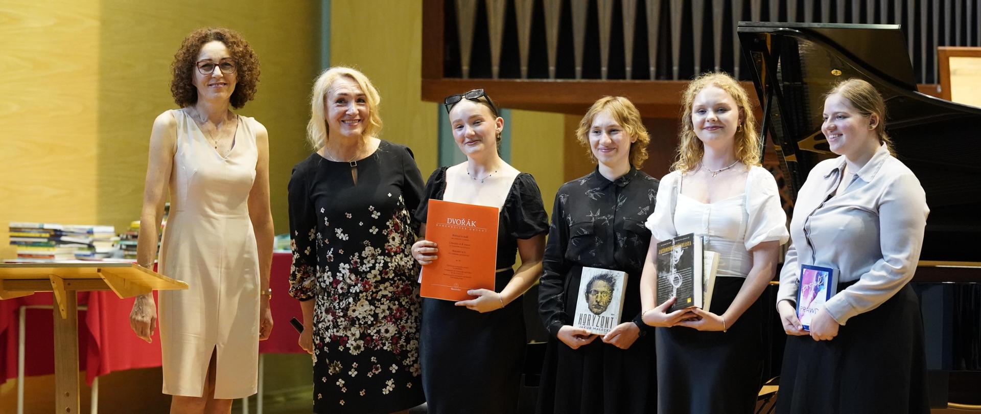 Na scenie sali koncertowej w jednym rzędzie pani dyrektor i wice stoją z absolwentkami II stopnia. Absolwentki trzymają w dłoniach nagrody książkowe. Za nimi fortepian oraz organy. Z lewej strony pod ścianą ułożone książki na ławkach.
