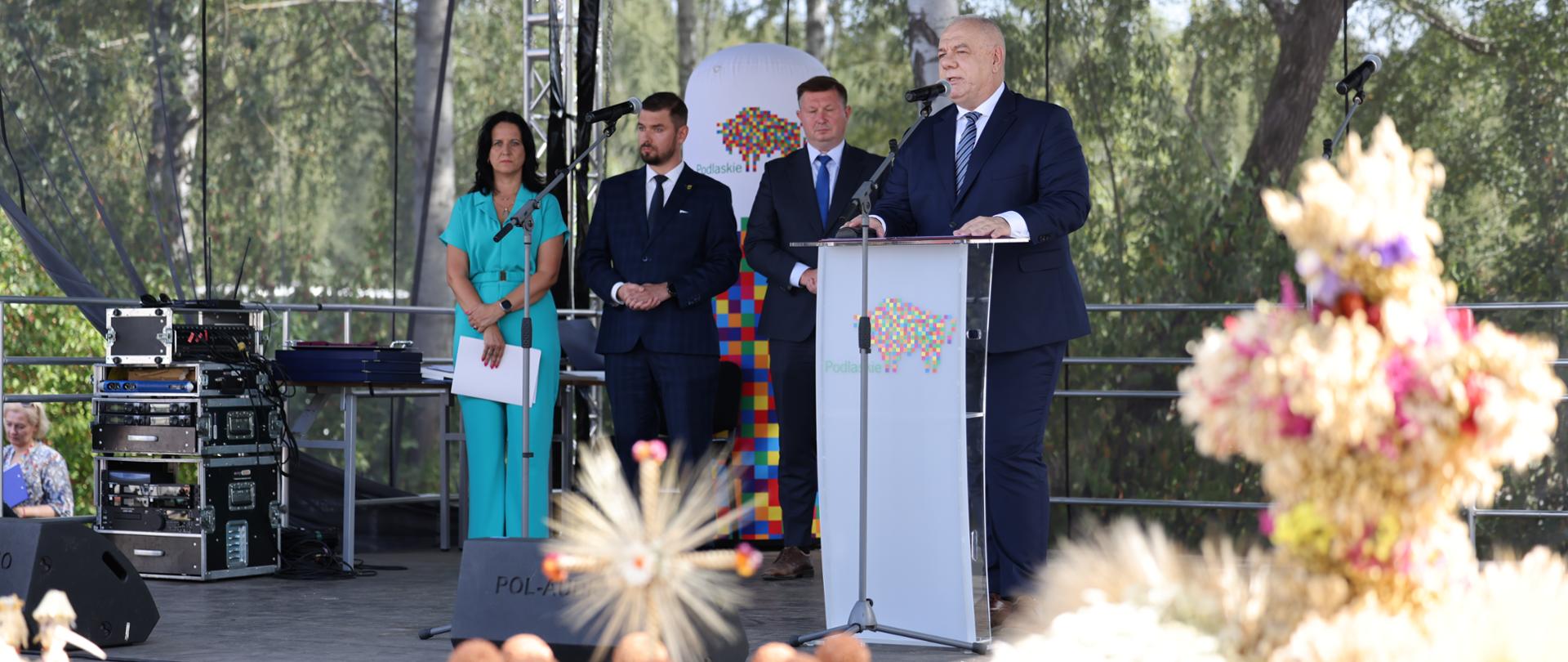 Minister Jacek Sasin stoi za mównicą. W tle stoi kobieta i dwóch mężczyzn.