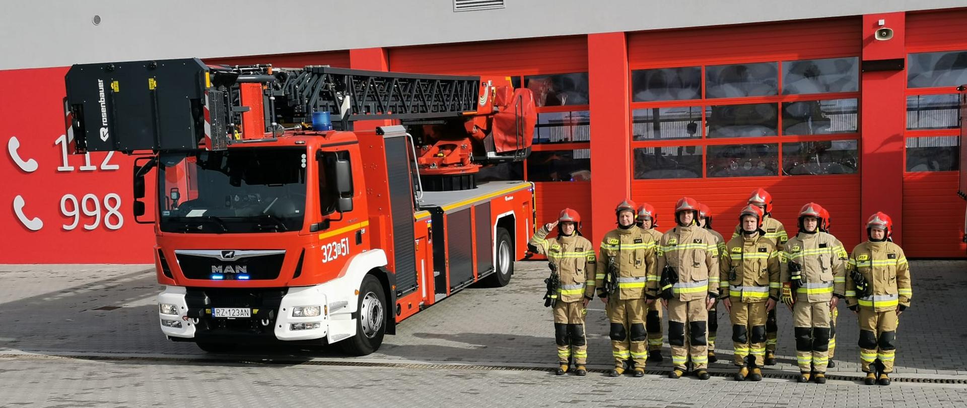 Zdjęcie przedstawia jedenastu strażaków w umundurowaniu bojowym, stojących przed garażem jednostki ratowniczo-gaśniczej w postawie zasadniczej. Obok ratowników wystawiono wozów - drabinę mechaniczną.