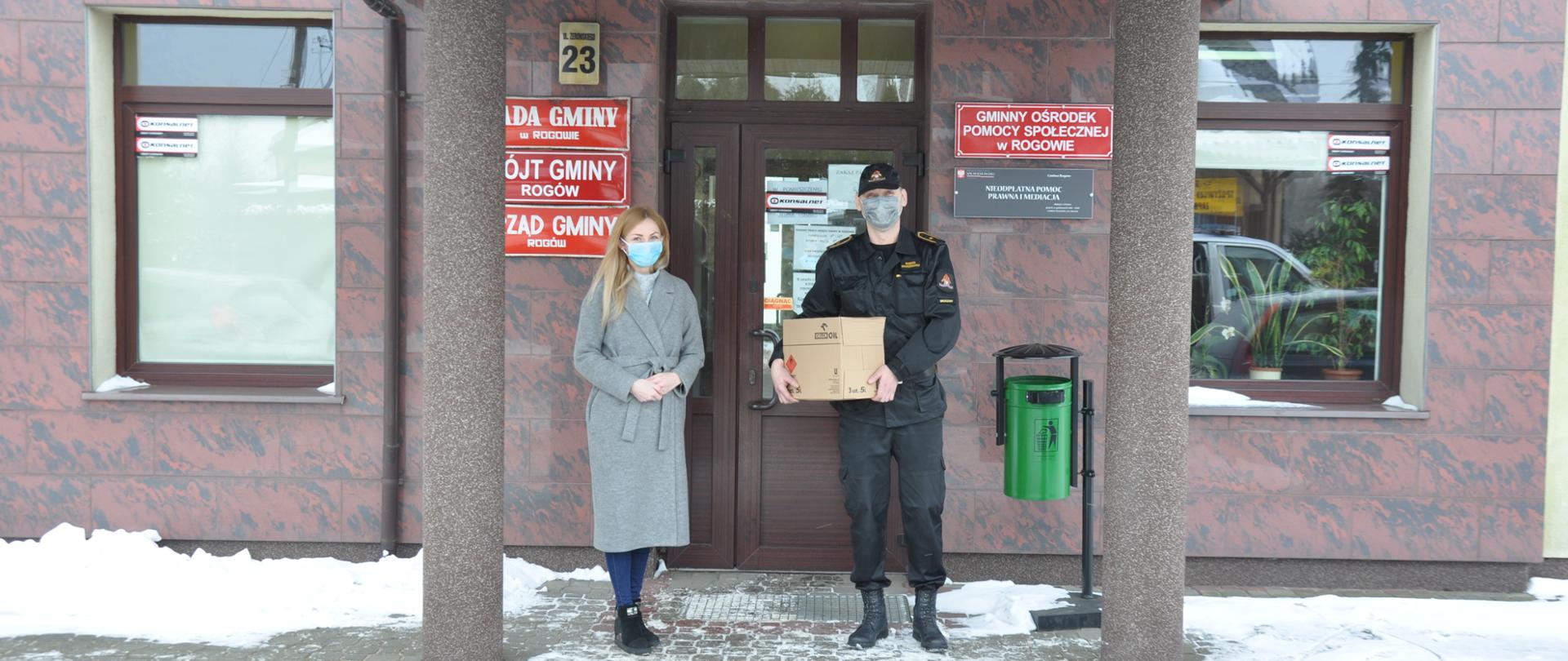 Zdjęcie zrobione na tle budynku Urzędu Gminy w Rogowie. Funkcjonariusz KP PSP w Brzezinach asp. Bogdan Banaszkiewicz przekazuje środki dezynfekcyjne przedstawicielce urzędu.