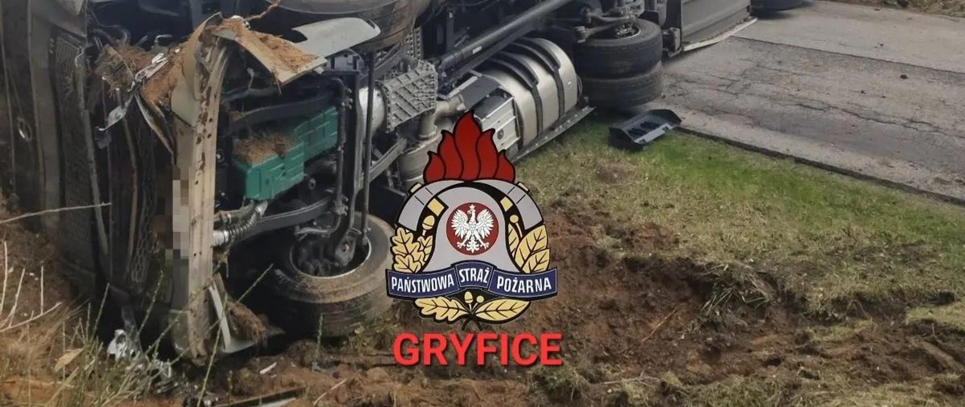 Na zdjęciu widoczny samochód ciężarowy z naczepą leżący na boku i blokujący drogę w obu kierunkach