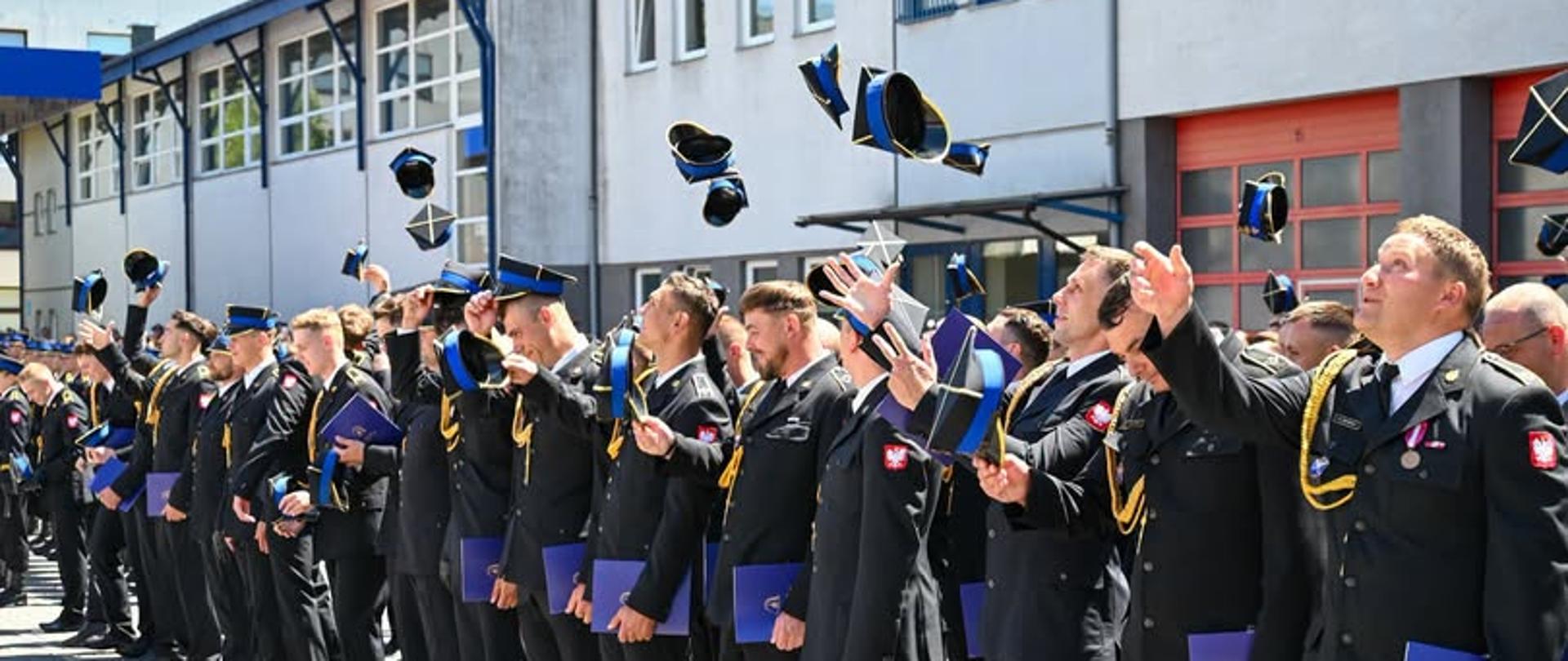 Strażacy w umundurowaniu galowym podczas promocji stojący w szyku podrzucają do góry rogatywki