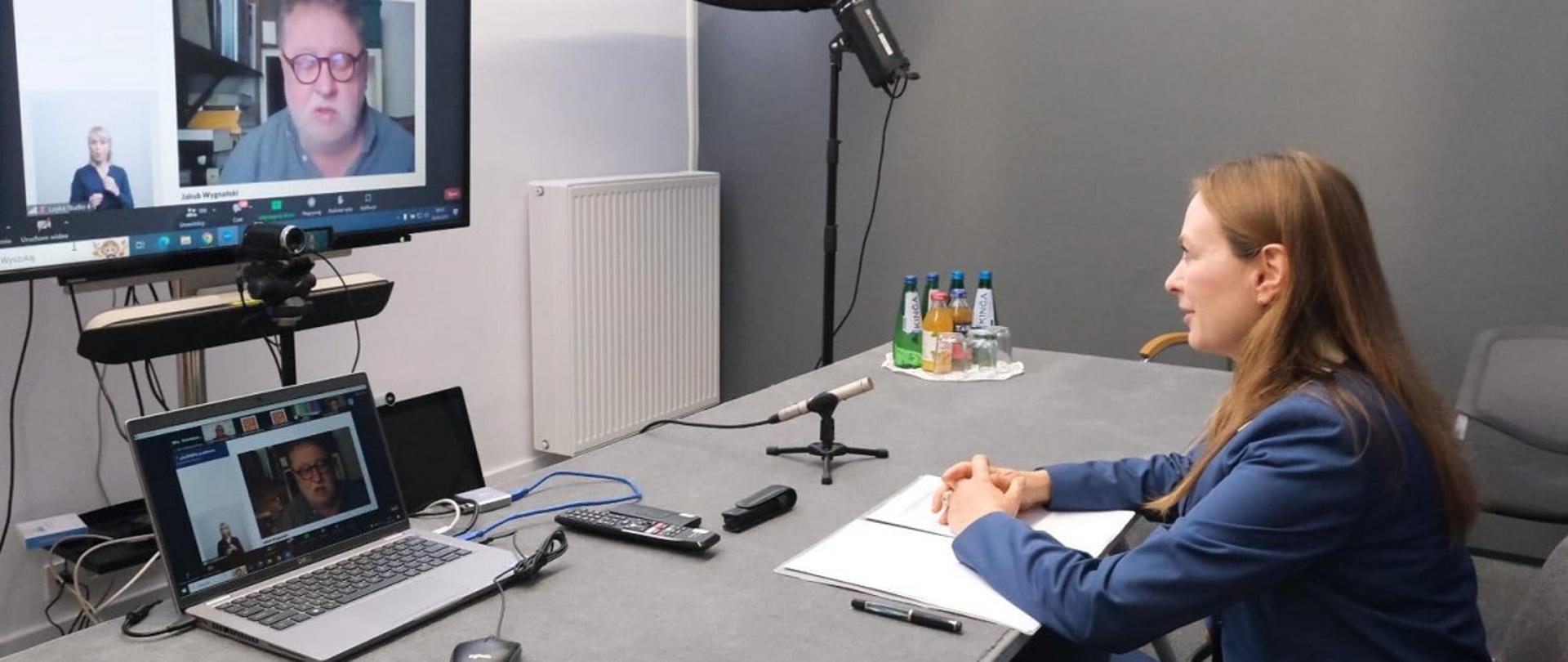 Minister funduszy i polityki regionalnej Katarzyna Pełczyńska-Nałęcz siedzi przy stole, przed nią laptop na którego ekranie widać mężczyznę