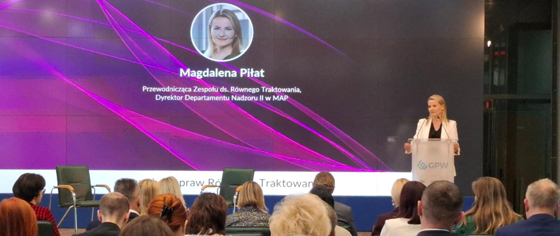 Uczestnicy spotkania siedzą na widowni. Przedstawicielka MAP przemawia przy mównicy. W tle ekran. 