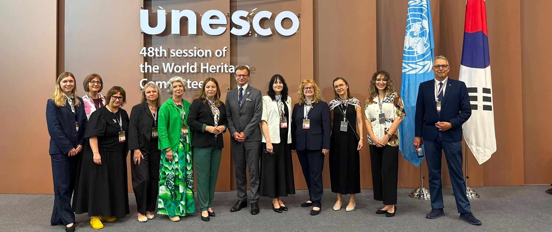 There are 12 people in the photo. Behind them is a brown background and the inscription: “UNESCO 48th session of the World Heritage Committee.” On the right are the flags of the United Nations and South Korea. 