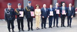 grupa ludzi stoi w szeregu, część z nich w mundurach strażackich, w środku grupy stoi kobieta w żółtym ubraniu