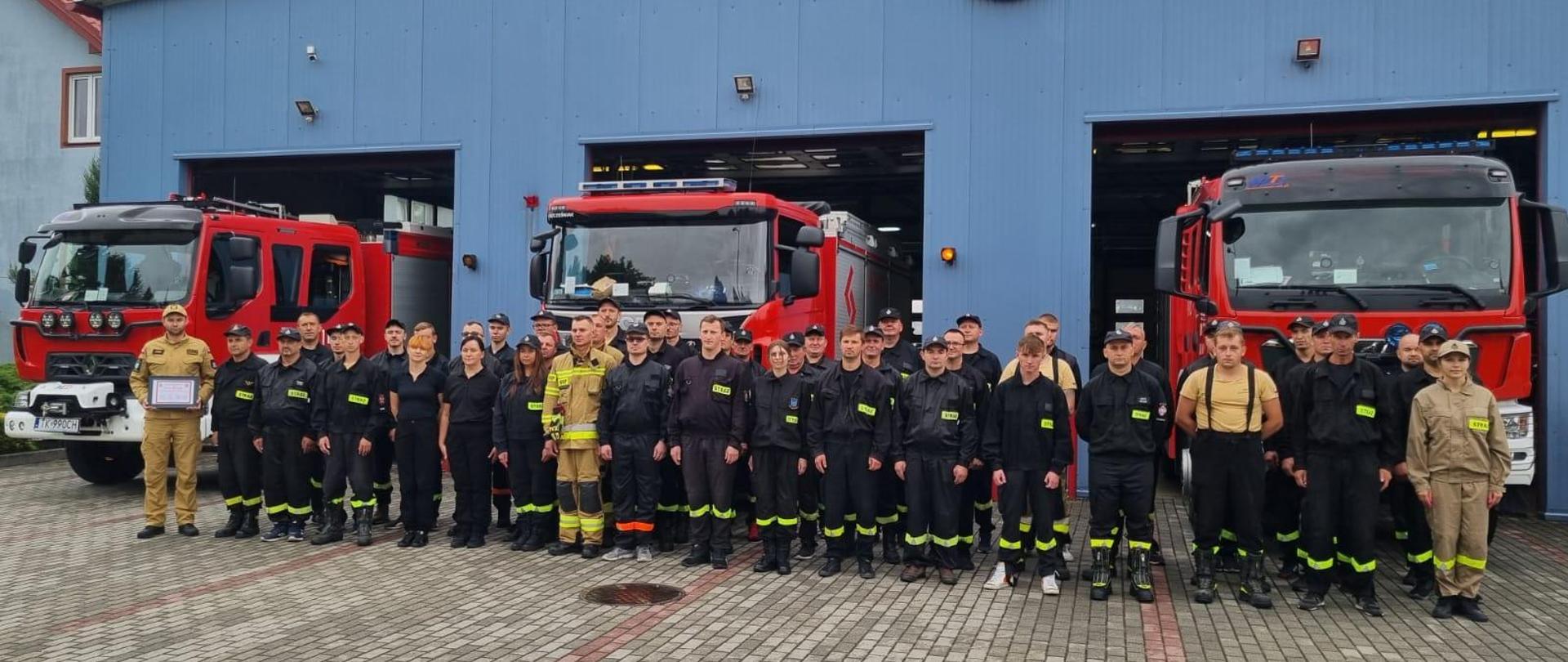Strażacy- ochotnicy w szyku ustawieni na tle wozów bojowych. 