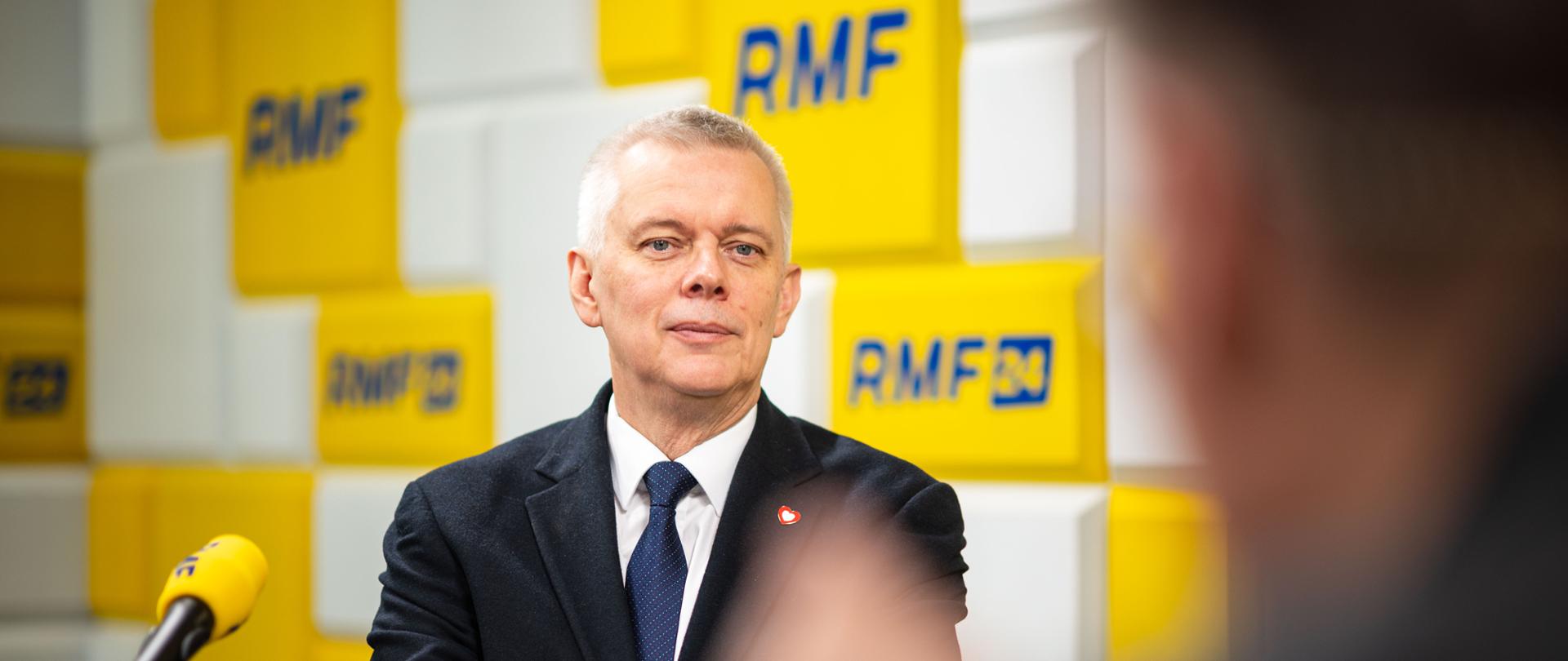 Minister Tomasz Siemoniak w garniturze i krawacie siedzi na tle żółtych prostokątów z logo RMF FM. W lewym dolnym rogu widoczny jest żółty mikrofon, a po prawej stronie rozmyta sylwetka innej osoby.