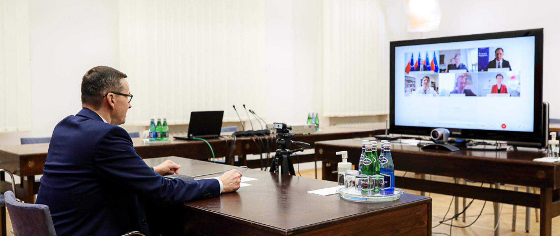 Premier Mateusz Morawiecki siedzi przy biurku, przed nim telewizor na którym widać innych uczestników wideokonferencji.