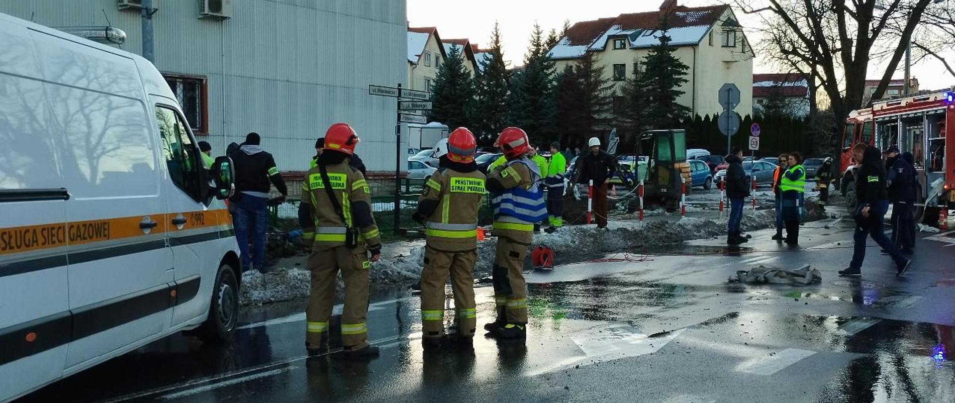 Na pierwszym planie stoi trzech strażaków PSP, w tym kierujący działaniem ratowniczym. Po lewej stronie samochód pogotowia gazowego. Po prawej funkcjonariusze policji, przedstawiciele zarządzania kryzysowego. Za nimi stoi samochód pożarniczy. 