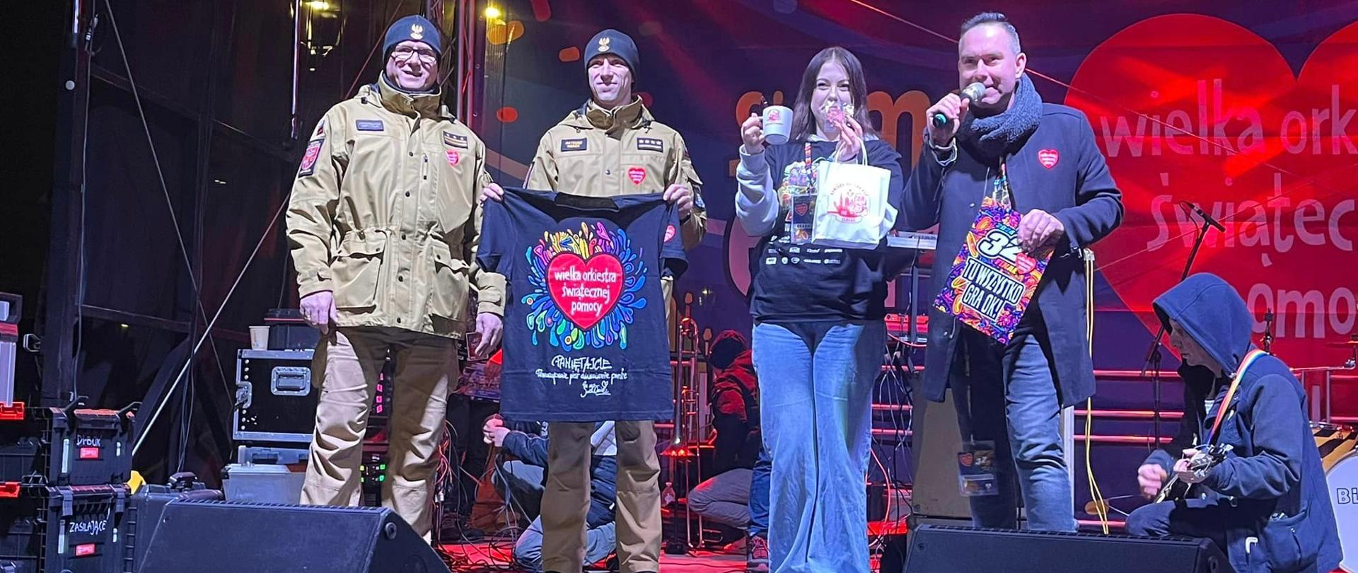 Na scenie stoi komendant elbląskich strażaków w towarzystwie innego strażaka w mundurze i pary prowadzącej imprezę. Strażacy trzymają w rękach koszulkę, która będzie licytowana. W tle duże czerwone serce z napisem Wielka Orkiestra Świątecznej Pomocy.