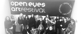 Grupa uczniów i nauczycielek stoi na klatce schodowej przed dużym banerem „Open Eyes Art Festival”. Wszyscy pozują do wspólnego zdjęcia.