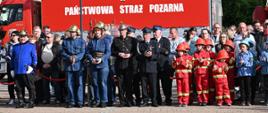 Uczestnicy apelu i obserwujący ludzie stoją na tle czerwonego samochodu pożarniczego z napisem na boku plandeki Panstwowa Straż Pożarna. W tle budynek.