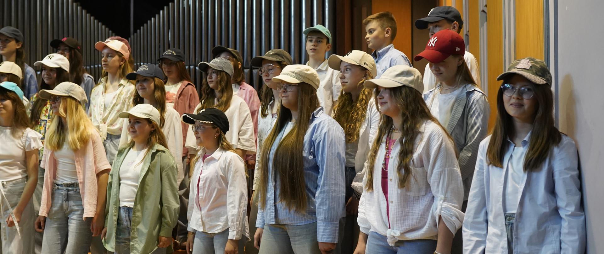 Na zdjęciu widoczna jest grupa młodych osób – dzieci i młodzieży – stojących na scenie w półokręgu, jak podczas występu chóralnego. Wszyscy mają na sobie codzienne, luźne stroje: jasne koszule, t-shirty, dżinsy i charakterystycznie – czapki z daszkiem w różnych kolorach. Scena znajduje się w sali koncertowej; w tle widać duże rury organów piszczałkowych. Panuje radosna, swobodna atmosfera – dzieci są skupione, jakby właśnie śpiewały lub przygotowywały się do rozpoczęcia występu. Na podłodze przed nimi leżą pluszowe zabawki, co może wskazywać na lekki, przyjazny charakter koncertu.